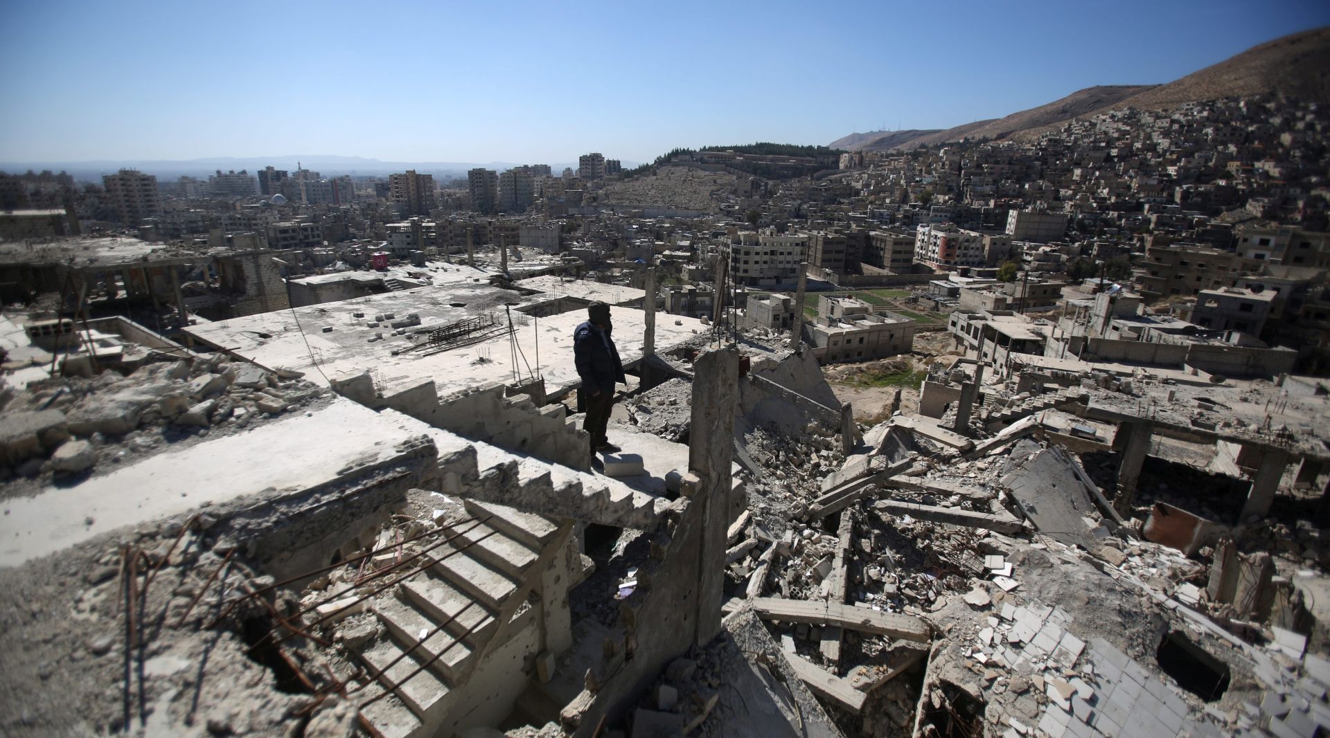 epa05166107 A Syrian man stands on top of a damaged building overlooking the Esh Al-Warwar neighborhood (background), in Barzeh neighborhood of Damascus, Syria, 17 February 2016. Barzeh neighborhood is controlled by rebels while the nearby Esh Al-Warwar is under control of Syrian government forces. A ceasefire is in effect between the two neighborhoods.  EPA/MOHAMMED BADRA