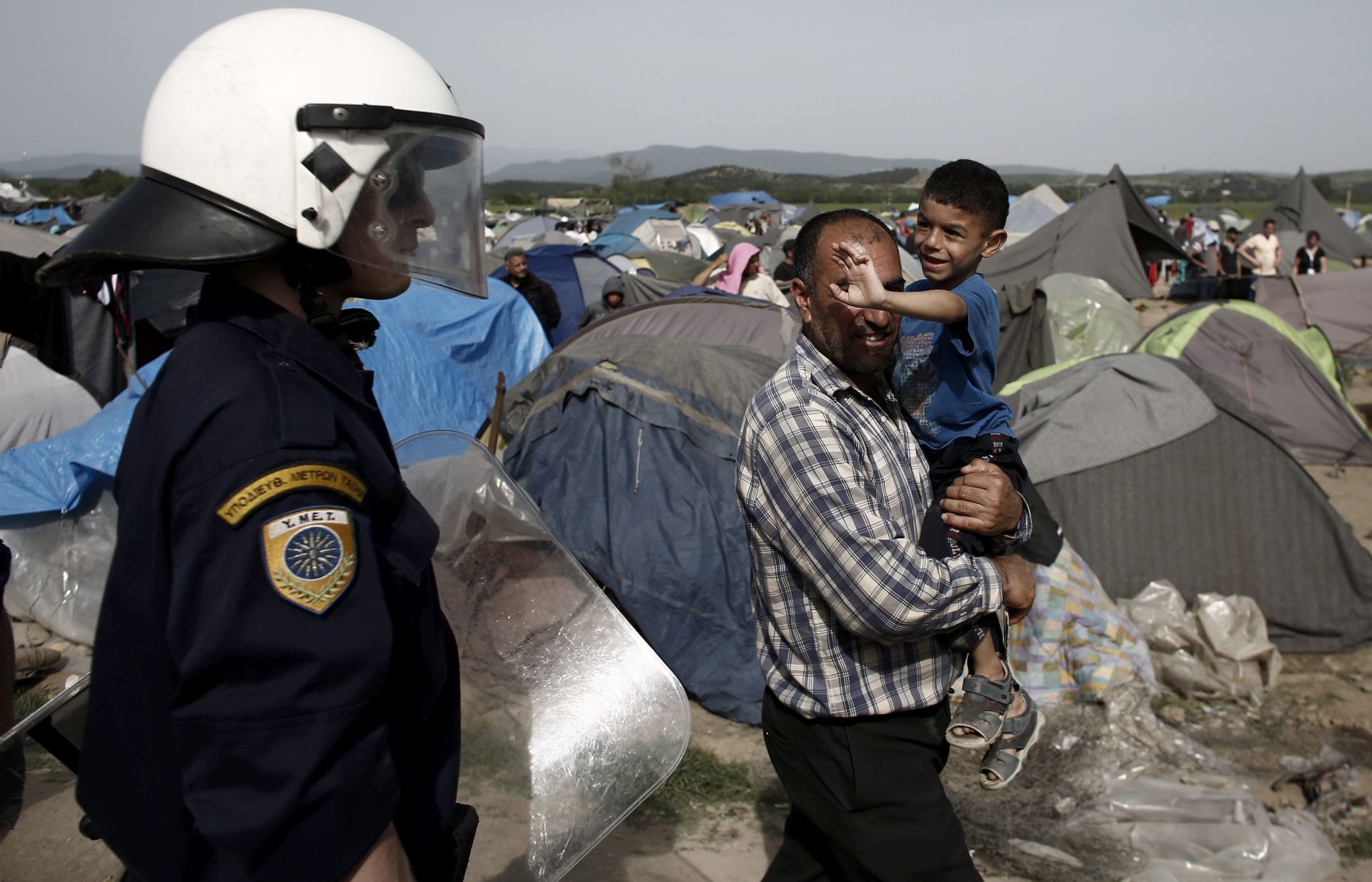 epa05265867 A refugee child waves to a Greek riot police officer after Greek riot police tried  to disperse refugees that threw rocks at a police van after a refugee was injured in the refugee camp of Idomeni near the Greek-FYROM borders, 18 April 2016.. A seriously injured refugee was rushed to a hospital from the camp at Idomeni, on the border between Greece and the Former Yugoslav Republic of Macedonia. The incident sparked tension in the camp, where residents attacked a police van that they considered was somehow involved, throwing stones and causing damage to the vehicle.  EPA/KOSTAS TSIRONIS