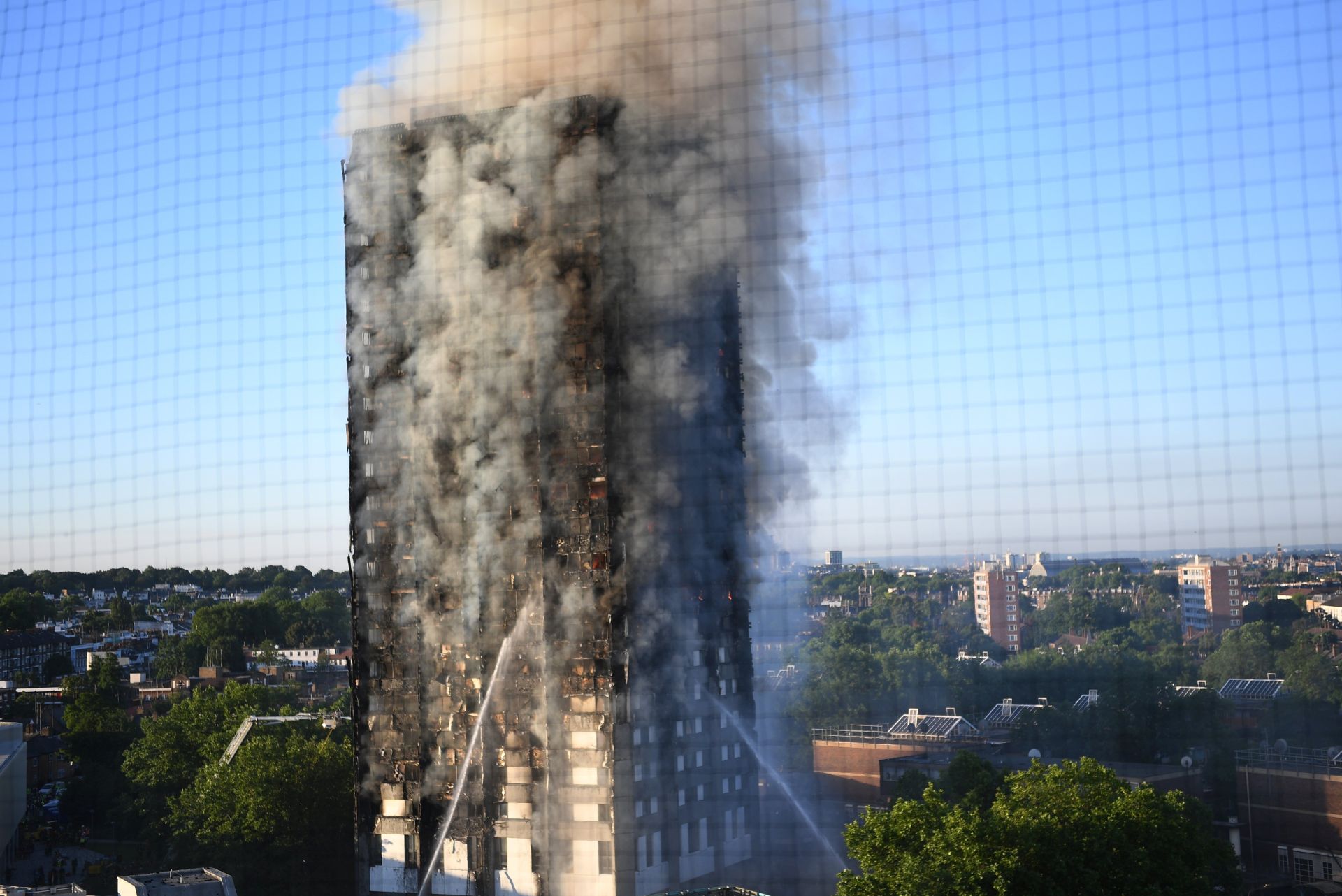 epa06027161 Smoke rises from the fire at the Grenfell Tower, a 24-storey apartment block seen through a fence in North Kensington, London, Britain, 14 June 2017. According to the London Fire Brigade, 40 fire engines and 200 firefighters are working to put out the blaze. Residents in the tower were said to be evacuating and a number of people were treated for a 'range of injuries,' Metropolitan Police said.  EPA/FACUNDO ARRIZABALAGA
