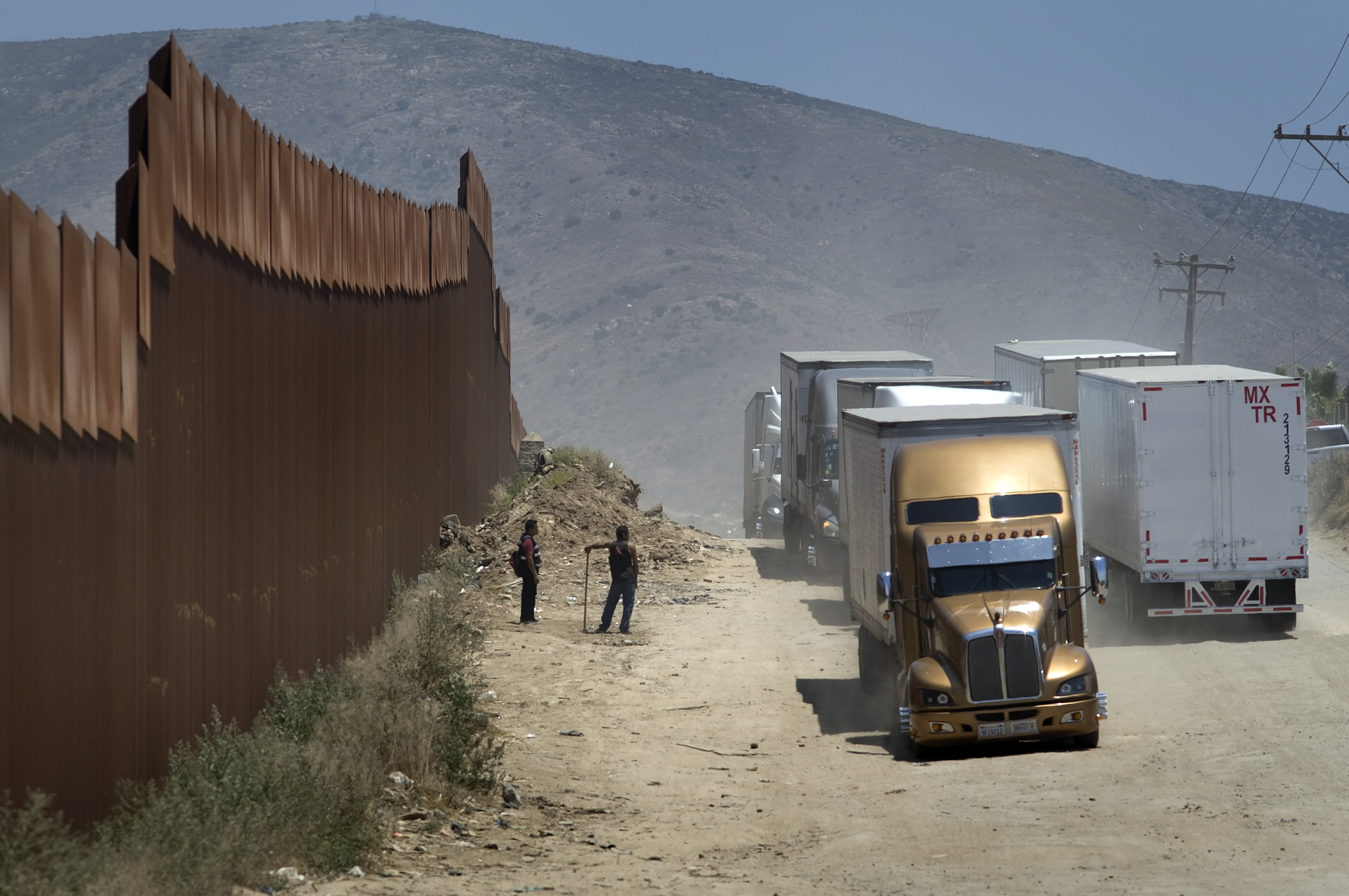 epa07626208 Two men who are fixing the road in exchange for tips watch as trucks drive along a dirt road next to the U.S.-Mexico border fence toward the commercial border inspection stationn in Tijuana, Mexico, on 04 June 2019, following US President Donald J. Trump's decision to impose tariffs on Mexico. The truck will queue in line with several hundred other trucks waiting to cross the border into San Diego, California, USA. US President Donald J. Trump stated he is planning to implement his decision to impose five percent tariffs on goods imported into the US from Mexico, with a five percent increase each month until October, if Mexican officials do not stop undocumented migrants from crossing into the US from the two nation's shared border.  EPA/David Maung