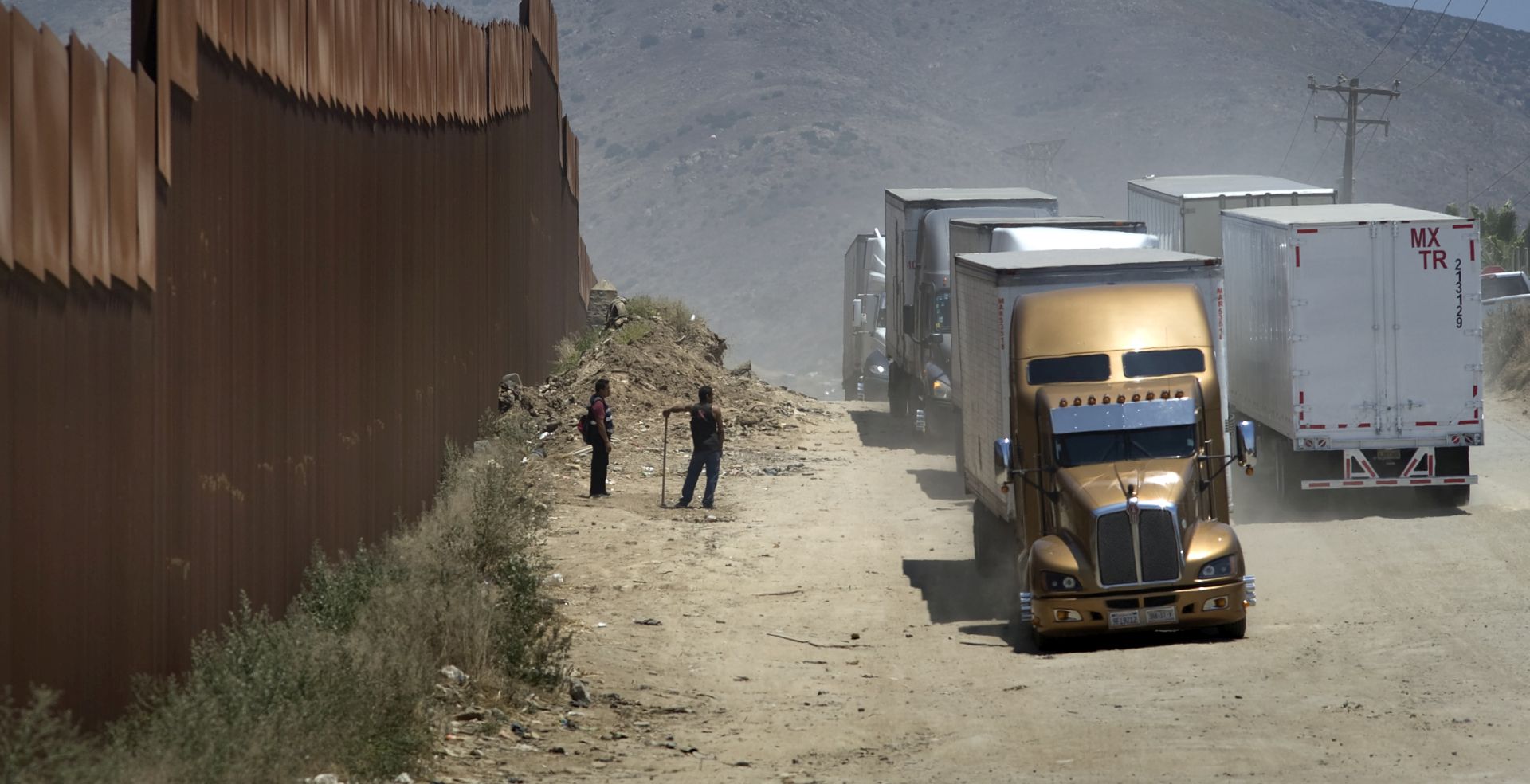 epa07626208 Two men who are fixing the road in exchange for tips watch as trucks drive along a dirt road next to the U.S.-Mexico border fence toward the commercial border inspection stationn in Tijuana, Mexico, on 04 June 2019, following US President Donald J. Trump's decision to impose tariffs on Mexico. The truck will queue in line with several hundred other trucks waiting to cross the border into San Diego, California, USA. US President Donald J. Trump stated he is planning to implement his decision to impose five percent tariffs on goods imported into the US from Mexico, with a five percent increase each month until October, if Mexican officials do not stop undocumented migrants from crossing into the US from the two nation's shared border.  EPA/David Maung