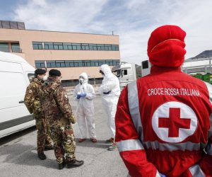 epa08322316 An Italian Red Cross member watches Italian soldiers speaking with employees in protective suits at the fruits and vegetables market of Fondi, near Rome, a farm town which has the largest wholesale produce market serving Rome and Naples, and has been put under even more stringent lockdown than the rest of the country due to a cluster of COVID-19 cases in Fondi, Italy, 25 March 2020.  EPA/GIUSEPPE LAMI