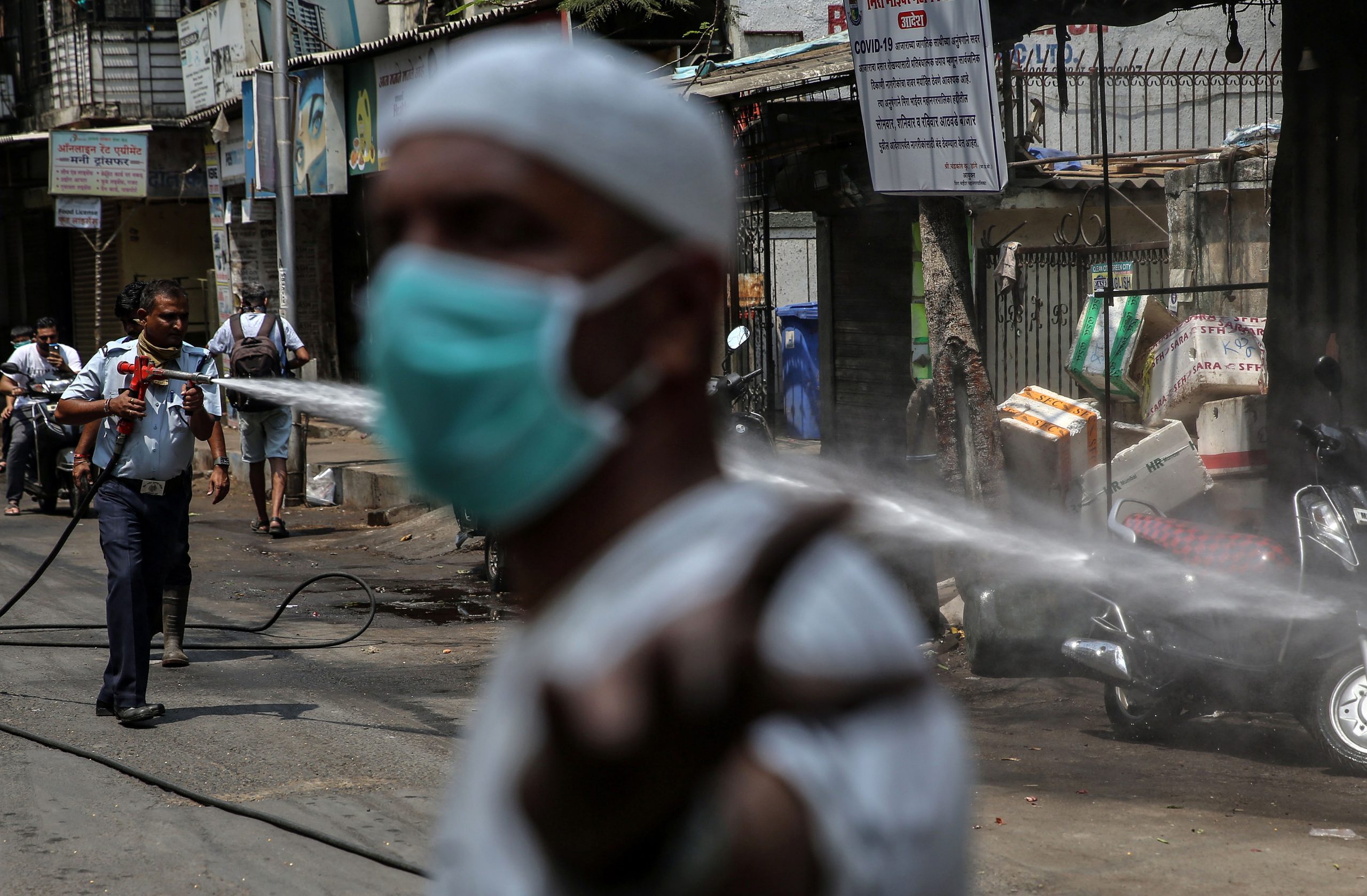 epa08323433 A man wearing a protective face mask walks in front of members of a municipal fire brigade as they spray disinfectant in a street  ofthe Mira Road residential area during an official lockdown of the city in an effort to combat the widespread of the SARS-CoV-2 coronavirus which causes the Covid-19 disease, in Mumbai, India, 26 March 2020. The Maharashtra state has been put under a  lock down until 31 March 2020 because of the Covid-19 coronavirus alert.  EPA/DIVYAKANT SOLANKI
