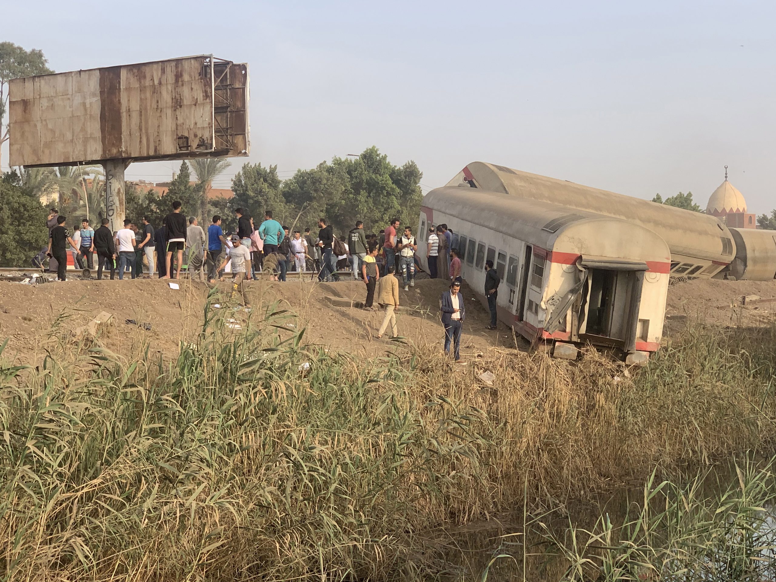 epa09143705 People stand near damaged carriages of a passenger train that was derailed in Toukh, Al Qalyubia Governorate, north of Cairo, Egypt, 18 April 2021. According to the ministry of health, 97 people were injured when several wagons of a passenger train that was heading to the city of Mansoura came off the rails.  EPA/KHALED ELFIQI