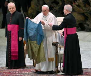epa09873205 Pope Francis, standing between Italian priest Monsignor Leonardo Sapienza (L) and Argentinian priest Monsignor Luis Maria Rodrigo Ewart (R), shows a national flag of Ukraine that was sent to him from the Ukrainian town of Bucha, during the weekly general audience in the Paul VI Audience Hall, in Vatican City, 06 April 2022.  EPA/ETTORE FERRARI