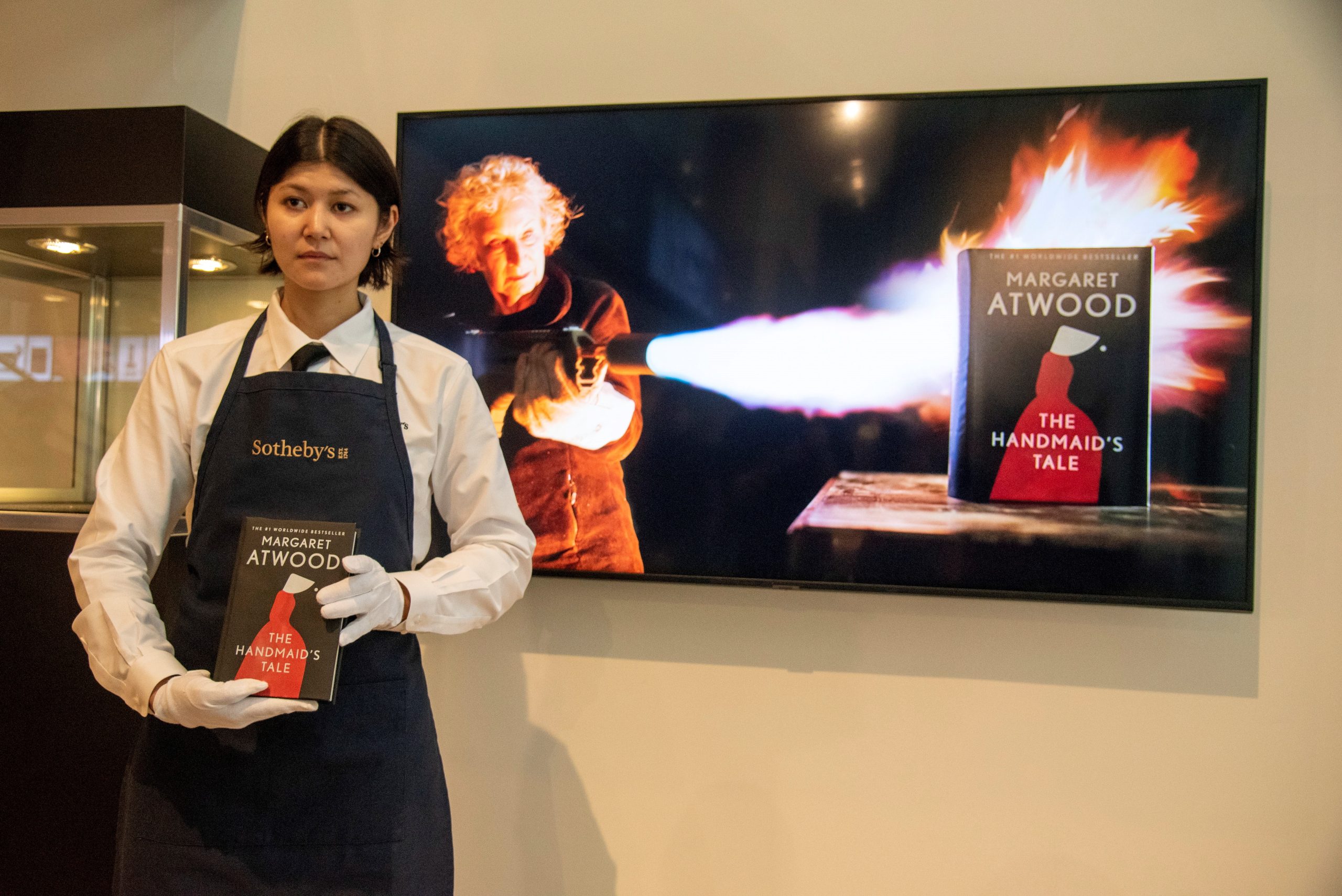 epa09993851 An art handler presents a fireproof edition of Margaret Atwood's 'The Handmaid's Tale' as a video of Atwood using a flamethrower plays on a screen behind them at the Sotheby's auction house in New York, New York, USA, 03 June 2022. Atwood and Penguin Random House partnered to make the special edition of Atwood's often banned book, up for auction online until June 7th.  EPA/SARAH YENESEL
