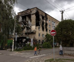 epa10080455 People walk past a building that was damaged during Russian attacks, in Hostomel, Ukraine, 19 July 2022. Hostomel, Irpin as well as other towns and villages in the northern part of the Kyiv region became battlefields and were heavily shelled when Russian troops tried to reach the Ukrainian capital Kyiv in February and March 2022. Russian troops on 24 February entered Ukrainian territory, starting a conflict that has provoked destruction and a humanitarian crisis.  EPA/ROMAN PILIPEY