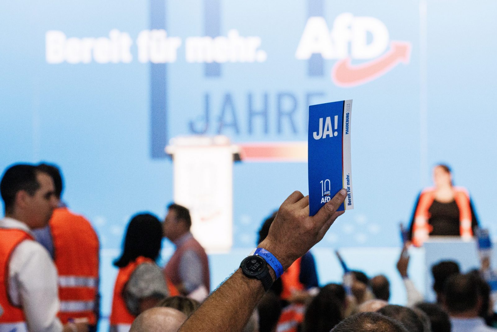 epa10773477 A delegate holds a voting card reading 'yes' as members of the counting committee walk in the background (orange vests) during the Alternative for Germany (AfD) party convention in Magdeburg, Germany, 28 July 2023. The Alternative for Germany (AfD) 14th federal party congress takes place in Magdeburg on 28 July, followed by a five-day European Elections assembly.  EPA/CLEMENS BILAN