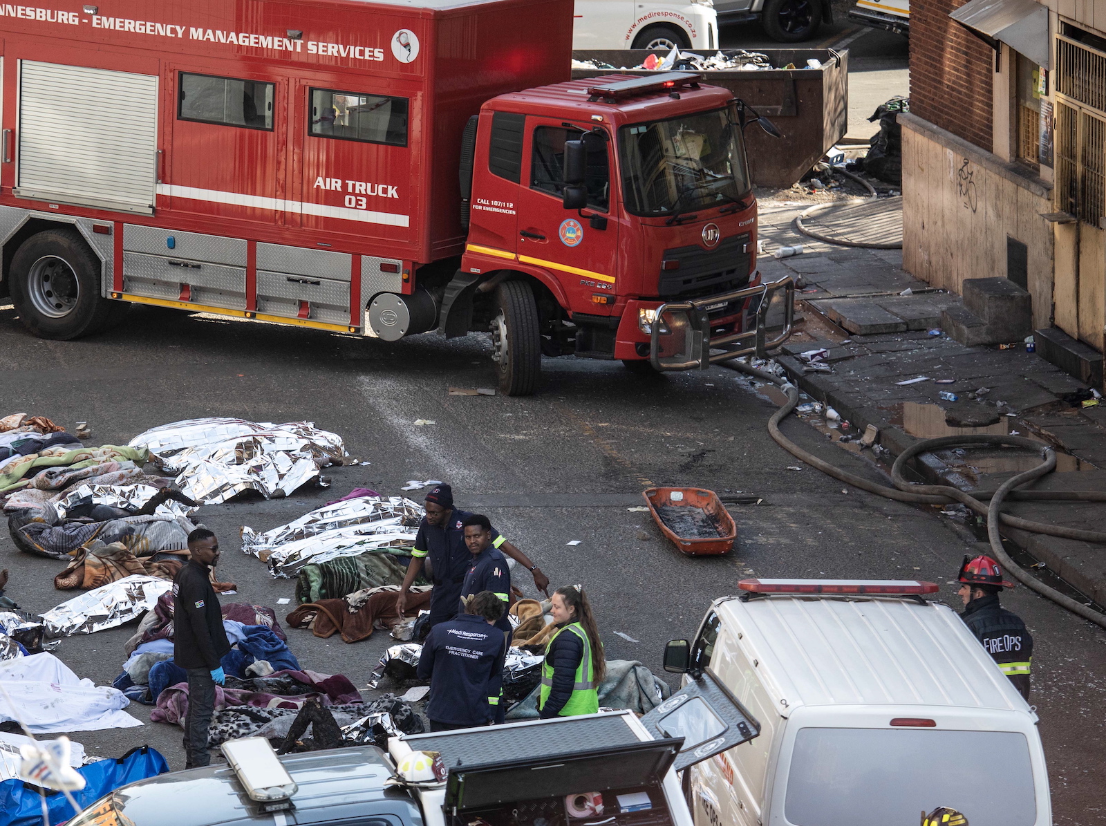 epa10830422 Fire crews and emergency staff stand next to the covered bodies of the victims as they gather at the site of a fire that broke out at the five-storey building in the city centre, in Johannesburg, South Africa, 31 August 2023. More than 60 people have died following a fire that ravaged a building in central Johannesburg according to officials.  EPA/KIM LUDBROOK
