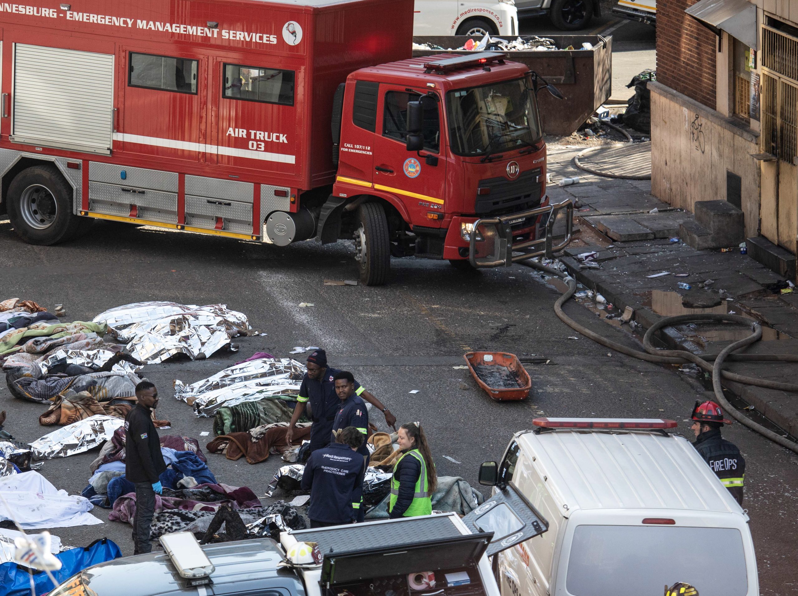 epa10830422 Fire crews and emergency staff stand next to the covered bodies of the victims as they gather at the site of a fire that broke out at the five-storey building in the city centre, in Johannesburg, South Africa, 31 August 2023. More than 60 people have died following a fire that ravaged a building in central Johannesburg according to officials.  EPA/KIM LUDBROOK