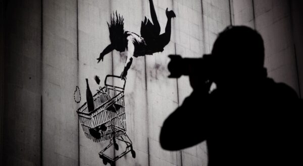 epa11003350 A person holds a camera to his face in front of a print of the artwork 'Falling Shopper' by Banksy during a press tour a the exhibition 'BANKSY - A Vandal Turned Idol' in Berlin, Germany, 30 November 2023. The unauthorized exhibition, showing original artwork and prints of British street art artist Banksy, is presented from 01 December 2023 at an unihabited property in Berlin.  EPA/CLEMENS BILAN