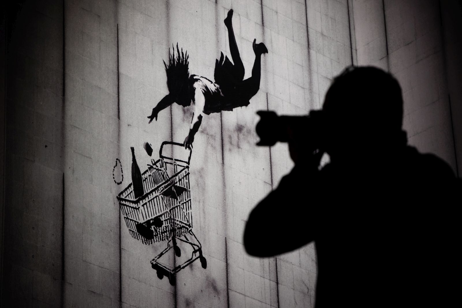 epa11003350 A person holds a camera to his face in front of a print of the artwork 'Falling Shopper' by Banksy during a press tour a the exhibition 'BANKSY - A Vandal Turned Idol' in Berlin, Germany, 30 November 2023. The unauthorized exhibition, showing original artwork and prints of British street art artist Banksy, is presented from 01 December 2023 at an unihabited property in Berlin.  EPA/CLEMENS BILAN